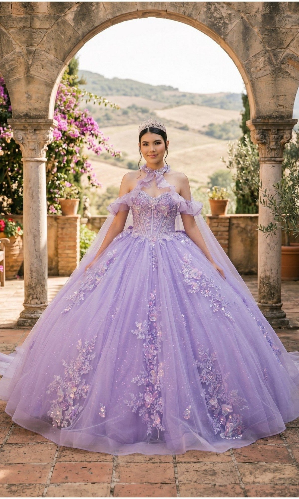 A young woman stands under a stone archway, wearing the Quinceanera Dress 8585JQ By Cinderella Couture in lilac, featuring a floral lace corset and delicate tiara, as the glitter tulle skirt shimmers while she poses outdoors with greenery and hills in the background.